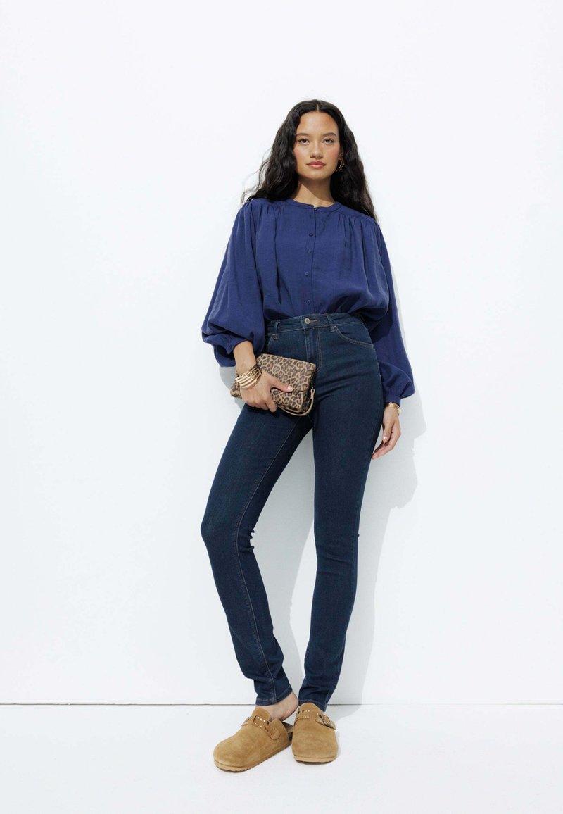 Blouse marine à manches bouffantes, jeans en denim foncé taille haute, et sabots en suède beige. Tenant une pochette en cuir imprimé léopard marron.