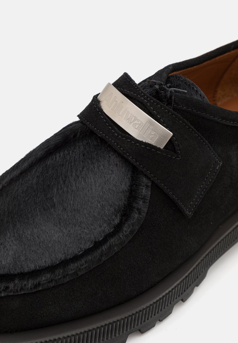 Chaussures noires alliant un mélange de daim lisse et de fourrure texturée, avec une sangle en métal argenté et un branding en relief. Semelle en caoutchouc avec des stries.