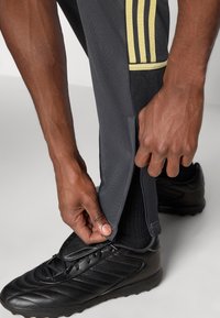 Mustad sportpüksid kollaste triipudega, kitseneva lõikega, reguleeritava lukuga pahkluul ja tekstureeritud kangaga. Osaliselt on nähtav must jalgpallijalats.