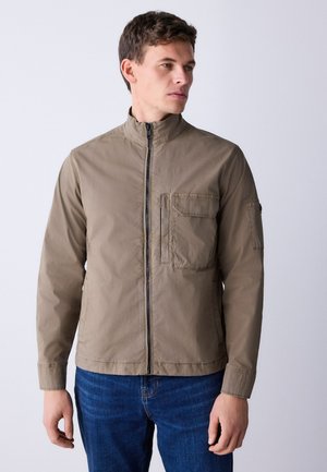 Mann trägt eine hellbraune Reißverschlussjacke mit Brust- und Ärmeltaschen, kombiniert mit blauen Jeans, blickt nach rechts vor schlichtem Hintergrund.