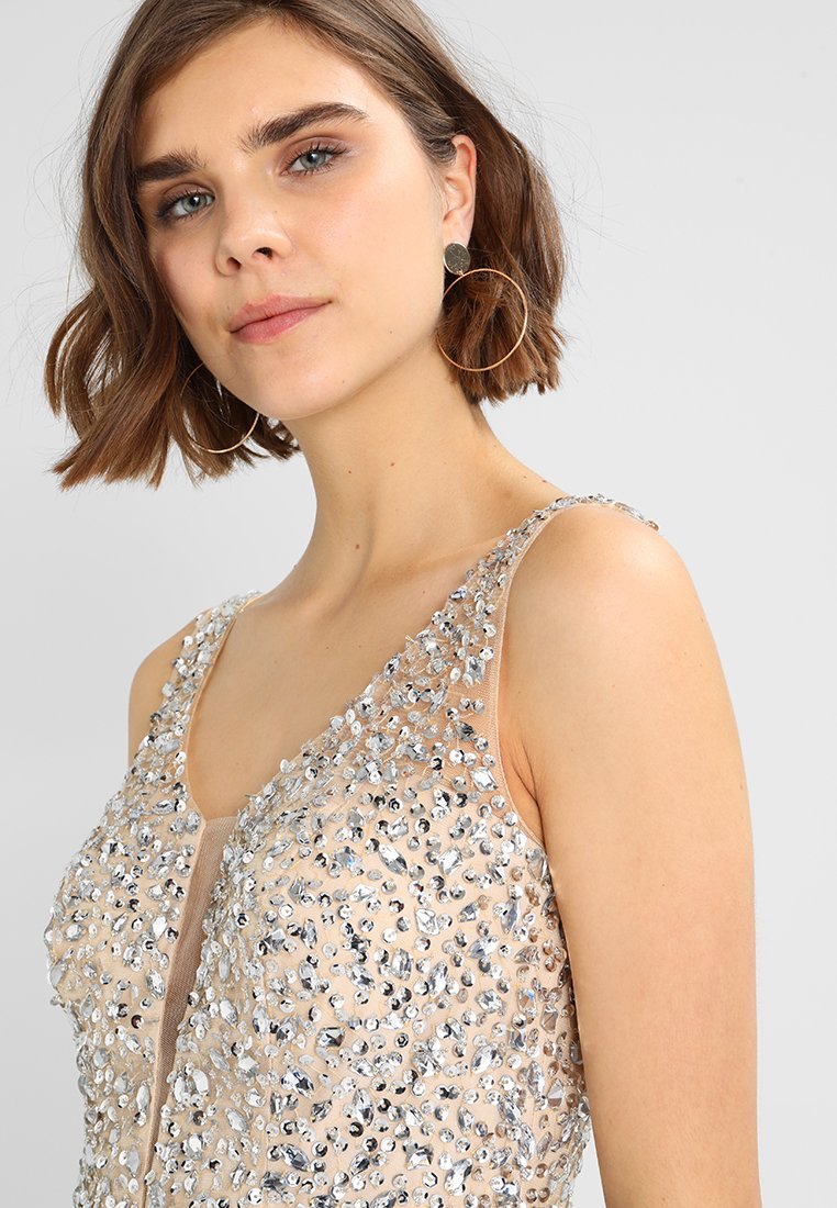 Beżowa sukienka z srebrnymi cekinami pokrywającymi górę, z dekoltem w kształcie litery V i cienkimi ramiączkami. Modelka nosi duże kolczyki w kształcie kół.