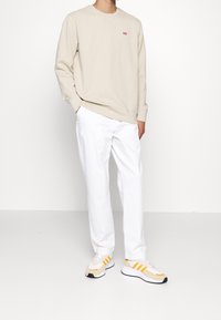 Felpa beige con collo rotondo e piccolo logo rosso, abbinata a pantaloni bianchi e sneakers beige con dettagli arancioni e neri.