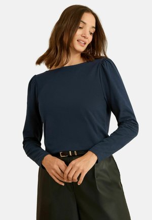 Naine, kes kannab tumesinist pikkade varrukatega särki ja musti pükse, käed sõrmedega kokku pandud, vaatab veidi alla neutraalse ilmega.