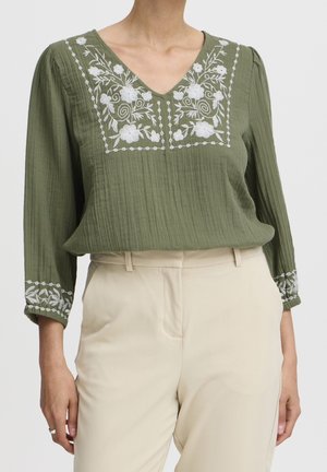 Olive green textured blouse with white floral embroidery on chest and cuffs, paired with high-waist beige trousers.