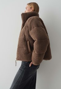 Braune Fleecejacke mit hohem Kragen, gepolstertem Design und einer Seitentasche. Verfügt über einen silbernen Reißverschluss und eine weiche, strukturierte Oberfläche.