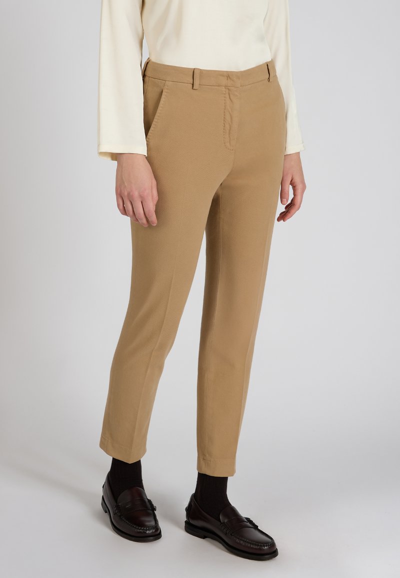 Jasnobrązowe dopasowane spodnie o gładkiej fakturze, z przednimi kieszeniami i dopasowanym kształtem. Połączone z ciemnymi mokasynami i widocznymi skarpetkami za kostkę.
