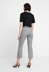 Kvinna i svart kortärmad topp, höga grå rutiga cropped byxor och svarta klacksandaler, som står mot vit bakgrund.