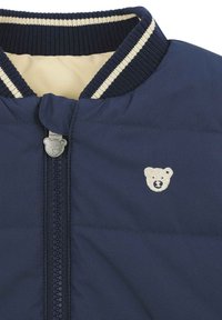 Giacca blu navy con colletto a coste con righe crema, emblema di un orso e una cerniera con un tirante a forma di orso. Texture liscia.