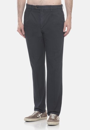 Uomo che indossa pantaloni dritti grigio scuro e sneakers beige, in piedi davanti a uno sfondo bianco semplice.