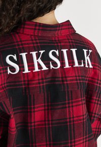 Camicia di flanella a quadri rossi e neri con "SIKSILK" ricamato in bianco sulla schiena. Presenta un colletto standard e un design con bottoni.