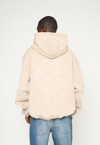 Bežová mikina s veľkým bielym grafikou na chrbte, ktorá zobrazuje "MULTIPLY APPAREL." Mäkká textúra, nadrozmerný strih a kapucňový dizajn.
