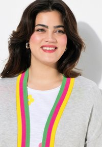 Lichtgrijze cardigan met meerkleurige gestreepte afwerking in groen, roze en geel, gedragen over een wit shirt met bloemdesigns.