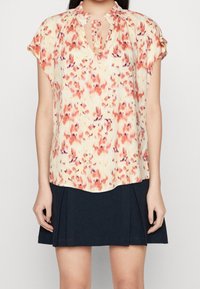 Blusa floreale nei toni del giallo pastello, rosa e viola, con scollatura a occhiello e maniche corte plissettate. Abbinata a una gonna plissettata scura.