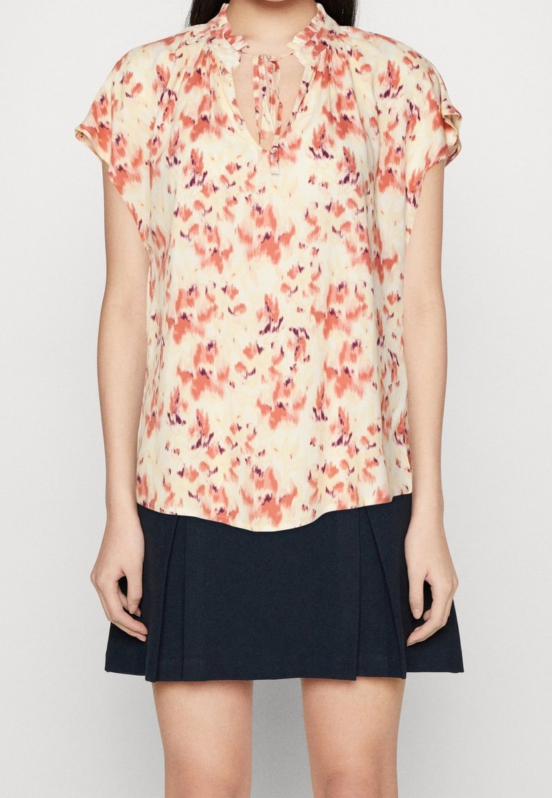 Blusa floreale nei toni del giallo pastello, rosa e viola, con scollatura a occhiello e maniche corte plissettate. Abbinata a una gonna plissettata scura.