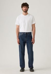Hombre adulto con cabello rizado, vistiendo una camiseta blanca lisa, jeans rectos azul y zapatos negros, de pie contra un fondo neutro.