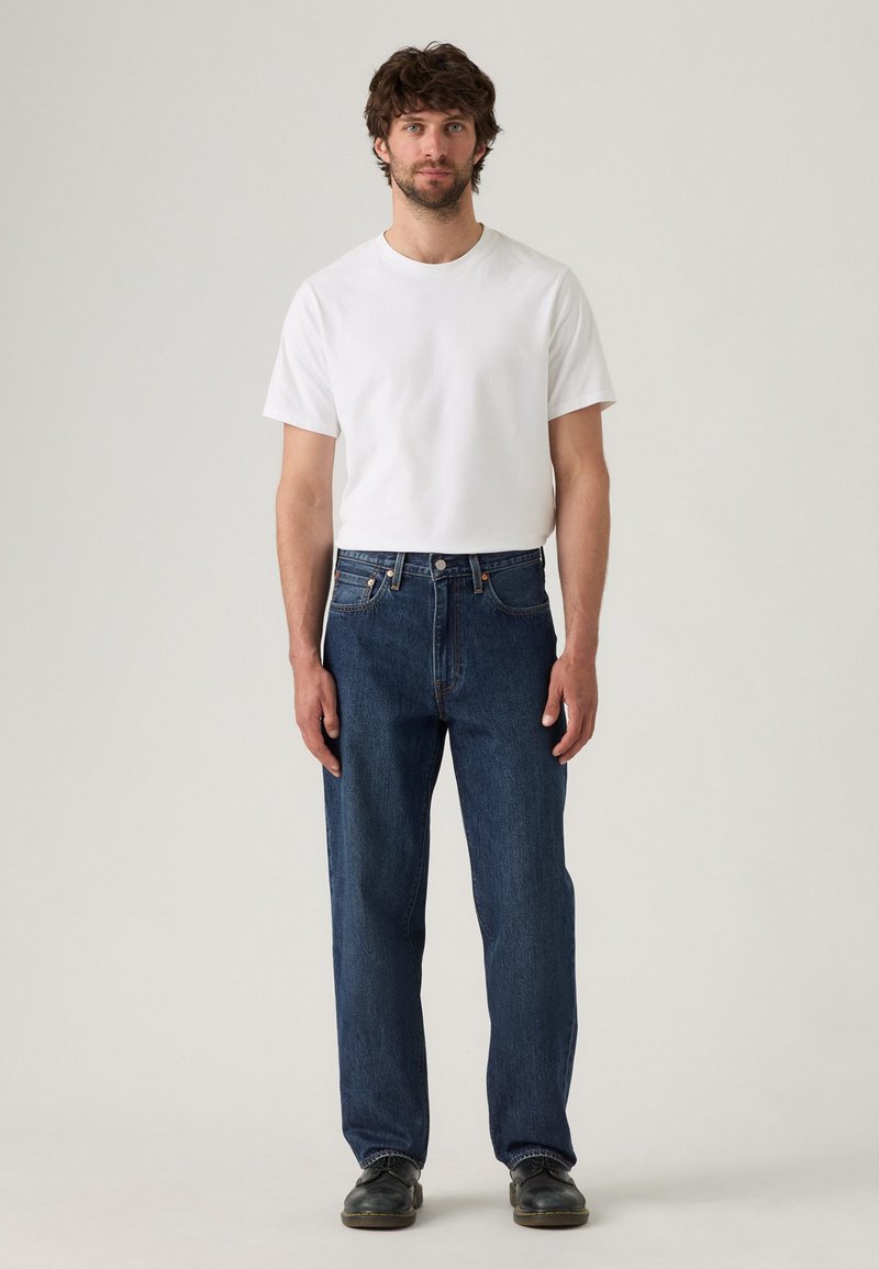 Hombre adulto con cabello rizado, vistiendo una camiseta blanca lisa, jeans rectos azul y zapatos negros, de pie contra un fondo neutro.