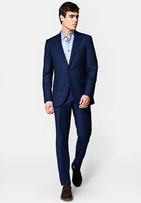 Granatowy garnitur w slim fit, z klapami i zapięciem na dwa guziki. Pod spodem jasnoniebieska koszula w wzory, w zestawieniu z ciemnymi butami.