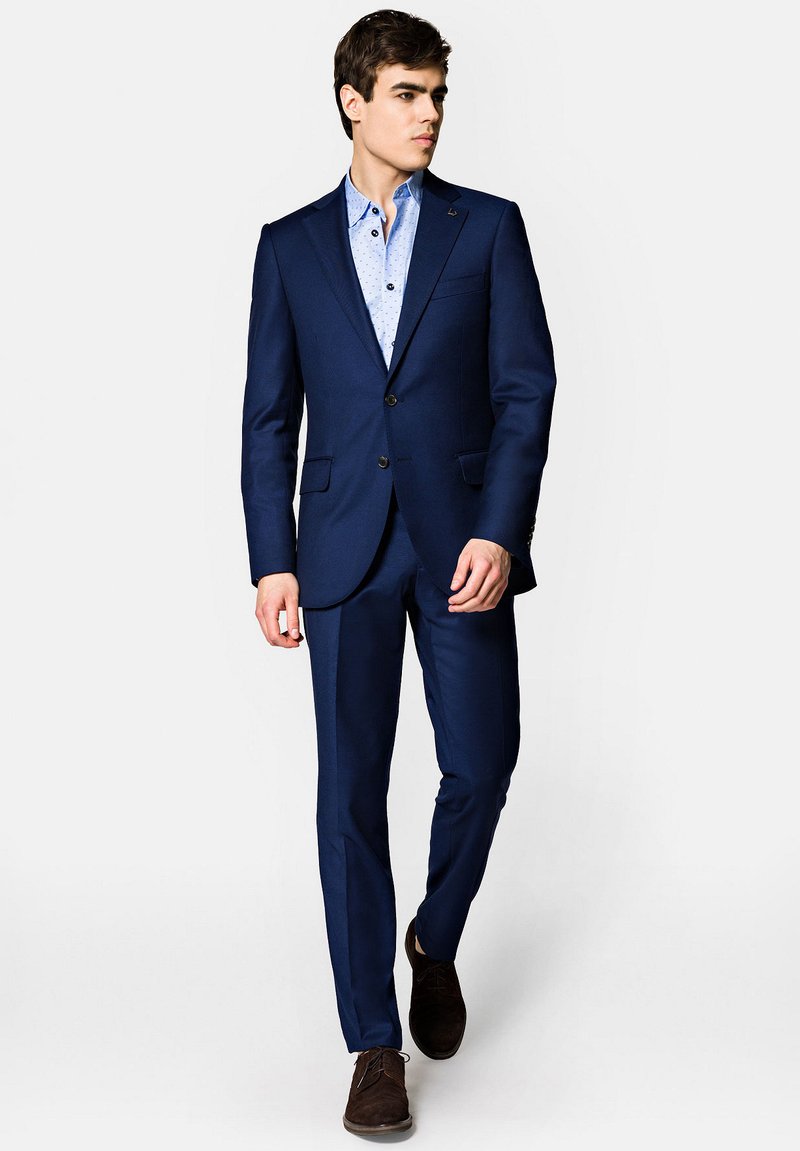 Granatowy garnitur w slim fit, z klapami i zapięciem na dwa guziki. Pod spodem jasnoniebieska koszula w wzory, w zestawieniu z ciemnymi butami.