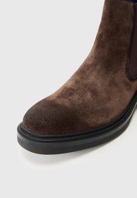 Stivale alla caviglia in suede marrone con punta arrotondata, suola in gomma nera e pannelli laterali elastici. Presenta una texture liscia e una leggera brillantezza.