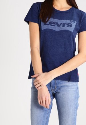 Vrouw die een marineblauw Levi's T-shirt en lichtblauwe spijkerbroek draagt, staand met armen gekruist bij de polsen voor een witte achtergrond.