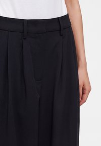 Gros plan d'une personne portant un pantalon plissé noir taille haute et une chemise blanche rentrée, la main droite détendue sur le côté.