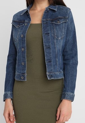 Frau trägt eine eng anliegende dunkelblaue Jeansjacke über einem olivgrünen, ärmellosen, gerippten Kleid und steht vor einem einfarbigen Hintergrund.