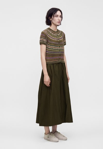 Femme portant un pull à manches courtes à motif, une longue jupe vert foncé et des baskets beige clair, debout devant un fond uni.