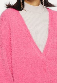 V-neck sweater in bright pink fuzzy knit, layered over a beige ribbed turtleneck. Distinctive soft texture and relaxed fit.