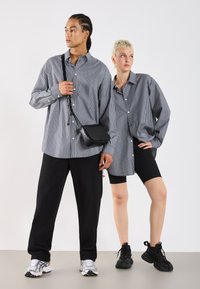 Två modeller poserar mot en vit bakgrund. Den ena har på sig en randig skjorta och håller en liten svart väska; den andra bär en lös skjorta och svarta shorts.