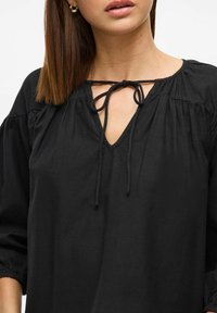 Melna blūze ar V veida izgriezumu un aizsienamu auklu, ar savelkamaām piedurknēm un mīkstu, vieglu audumu. Vienkāršs un brīvs dizains.