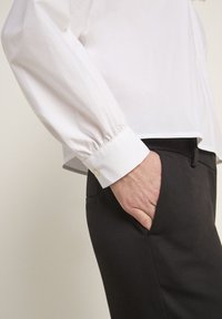 Weißes Langarmhemd mit gerafften Manschetten und einem Knopf. Kombiniert mit schwarzen Hosen mit Seitentasche. Glatte Stofftextur.