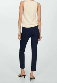 Donna in piedi di spalle, con un top senza maniche beige, pantaloni attillati blu navy e sandali neri con tacco, davanti a uno sfondo neutro.