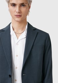 Mörkgrå blazer med strukturerade axlar, enkel knappstängning, slagkrage och bröstficka, bärs över en vit skjorta med knappar.