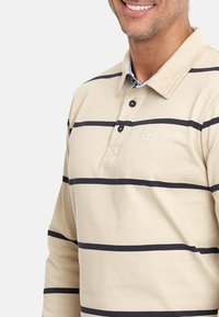 Camisa polo de manga larga color beige con franjas horizontales azul marino, que cuenta con un cuello, tapeta de botones y un pequeño logo bordado en el pecho.