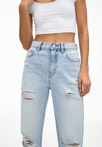 Jeans de cintura alta de color azul claro con parches desgastados y dobladillos deshilachados. Hechos de denim, cuentan con un diseño de cinco bolsillos y cierre de botón metálico.