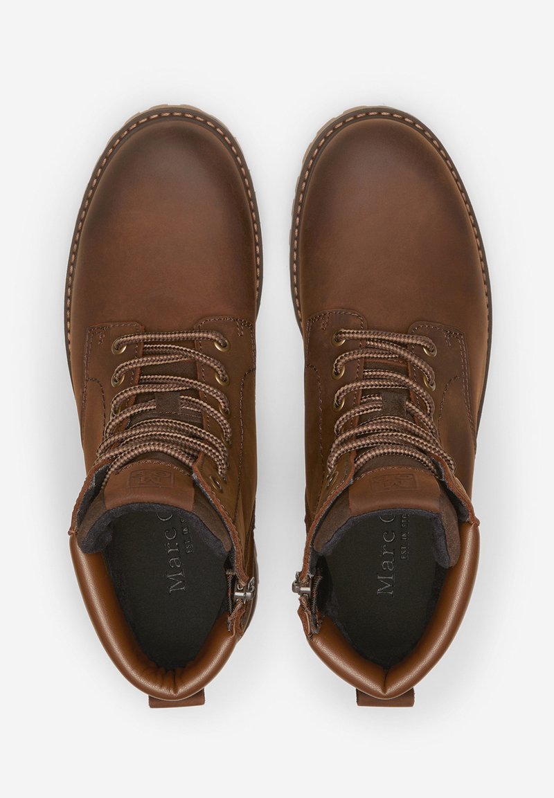 Botas de tobillo de cuero marrón con punteras redondas, ojales metálicos, diseño de cordones y forro acolchado. Un ligero pespunte realza los bordes exteriores.
