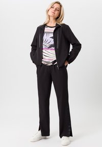 Zwarte ritssluiting jack met satijnen accenten, grafisch zwart-wit patroon shirt, zwarte broek met zijsplitten en witte sneakers.