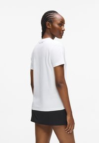 Valge puuvillane lühikeste varrukatega T-särk ümmarguse kaelusega. Seljal on kaeluse lähedal peen brändimine. Kombineeritud musta seelikuga, rõhutades mitmekülgsust.