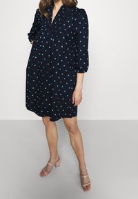 Mörkblå klänning med blåa prickar, placket-hals och puffärmar; i kombination med ljusrosa blockklack-sandaler mot en neutral bakgrund.