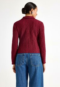 Bordeaux farvet tekstureret jakke med lange ærmer og krave, parret med blå denim jeans med baglommer.