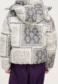 Prošívaná péřová bunda v šedé a béžové barvě s paisley a květinovými vzory. Má kapuci a je nošena přes fialové kalhoty.