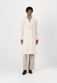 Hellbeige Mantel im Fischgrätmuster mit einem breiten Kragen, doppelten Knöpfen und langen Ärmeln, kombiniert mit cremefarbenen Hosen und schwarzen Stiefeletten.