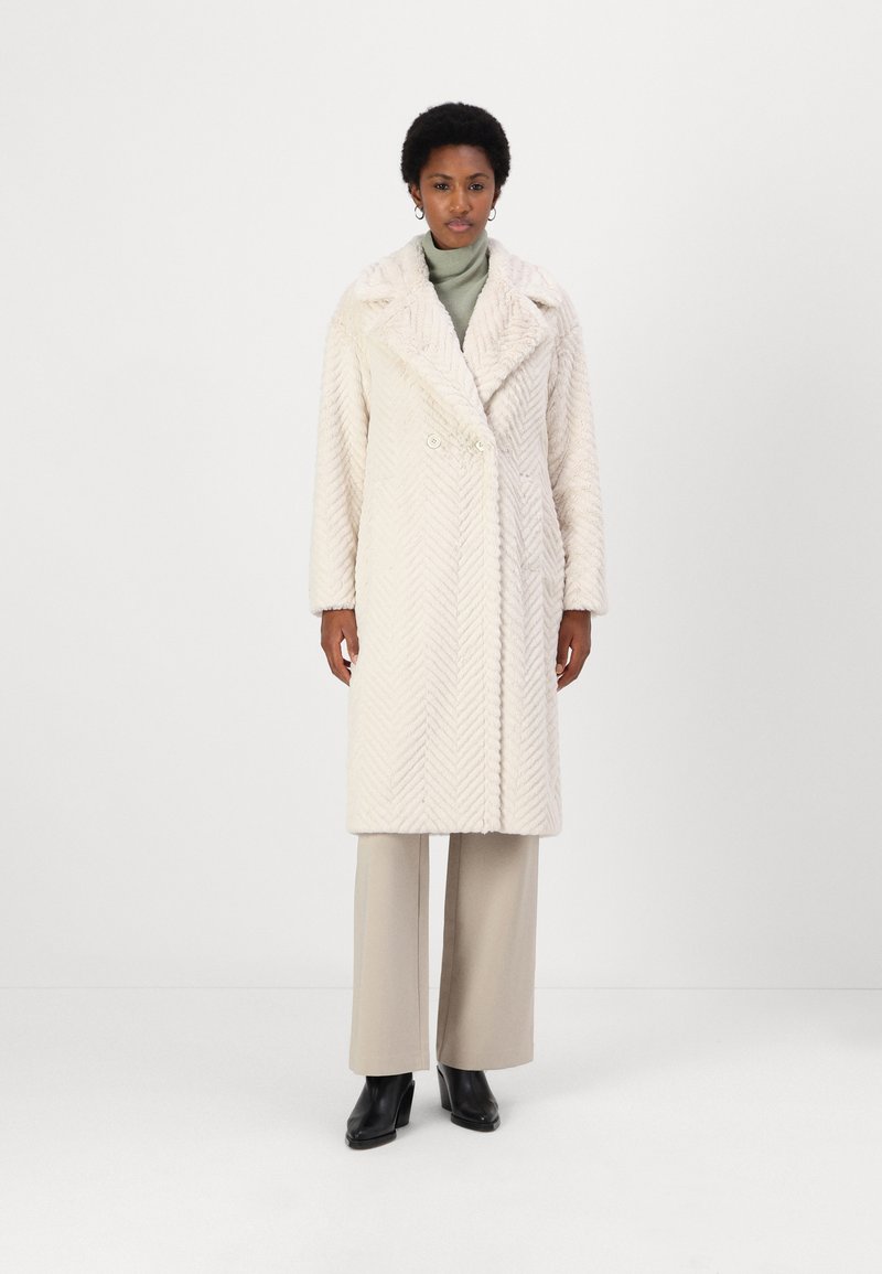 Hellbeige Mantel im Fischgrätmuster mit einem breiten Kragen, doppelten Knöpfen und langen Ärmeln, kombiniert mit cremefarbenen Hosen und schwarzen Stiefeletten.