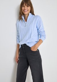 Hellblauer gestrickter Pullover mit halber Reißverschluss-Kragen, der Kabelstrickmuster aufweist, kombiniert mit dunkelgrauen, weiten Jeans und den Händen in den Taschen.