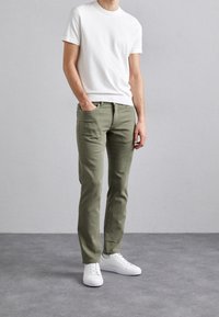 Biała koszulka z krótkim rękawem noszona z oliwkowozielonymi spodniami o slim-fit i białymi trampkami, stojąca na tle neutralnego szarego tła.