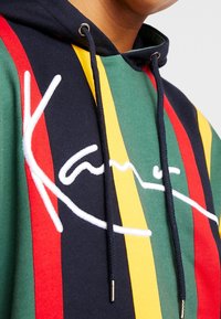 Acercamiento a una sudadera con rayas verticales en verde, amarillo, rojo y azul marino, que presenta un texto de firma bordado en blanco y cordones en azul marino.