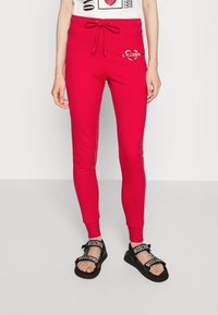 Czerwone dresy z elastycznym pasem i sznurkiem, z okrągłym logo "Love Moschino". Połączone z czarnymi sandałami z markowymi paskami.