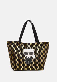 Bolsa tote negra con un patrón geométrico dorado y un personaje de dibujos animados con gafas de sol y traje en el frente. Asas negras.