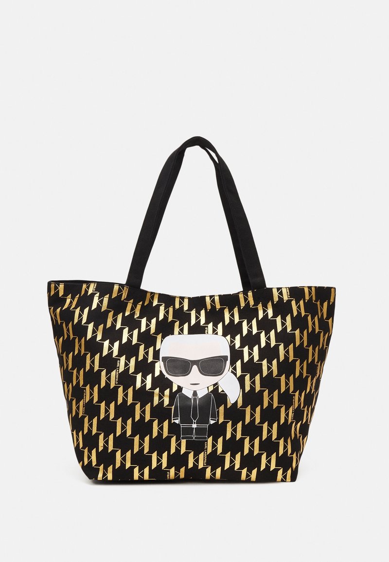 Bolsa tote negra con un patrón geométrico dorado y un personaje de dibujos animados con gafas de sol y traje en el frente. Asas negras.