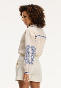 Blusa in tessuto crema con motivi ricamati blu sulle maniche e colletto, abbinata a pantaloni corti bianchi su misura.